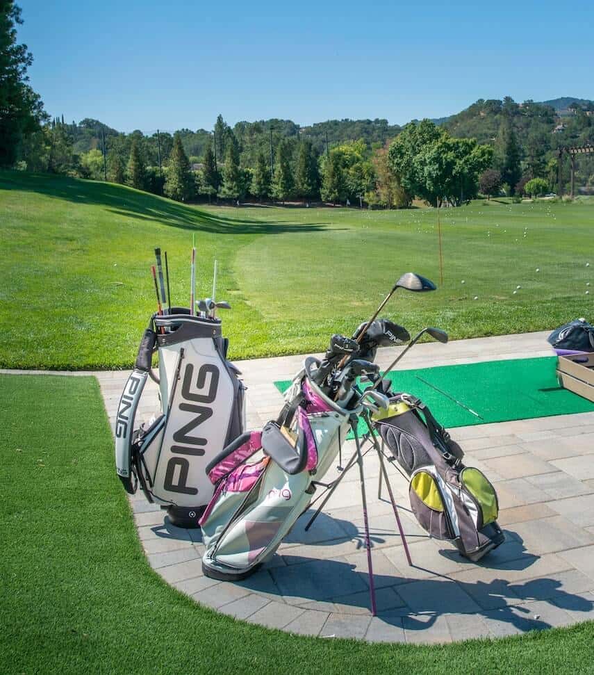 3 golf bags with clubs stood up on a tiled surface next to a golf course
