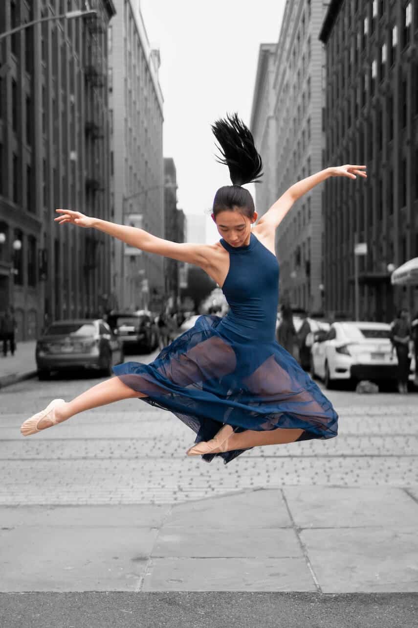 Ballerina in a blue leotard and chiffon skirt leaping on a bust street surrounded by high rise buildings