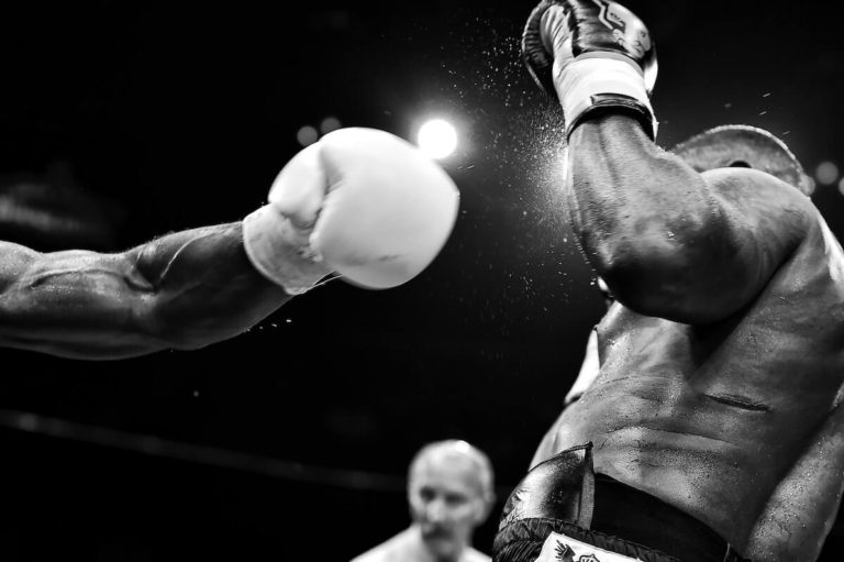 Boxing Quiz Questions and Answers cover photo of a man reeling from a punch from a man wearing a white boxing glove, the referee in a white shirt out of focus behind them both