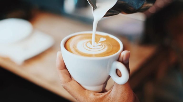 Coffee Trivia Questions and Answers cover photo of a hand holding a white cup of cofee that is having milk poured into it from a silver beaker