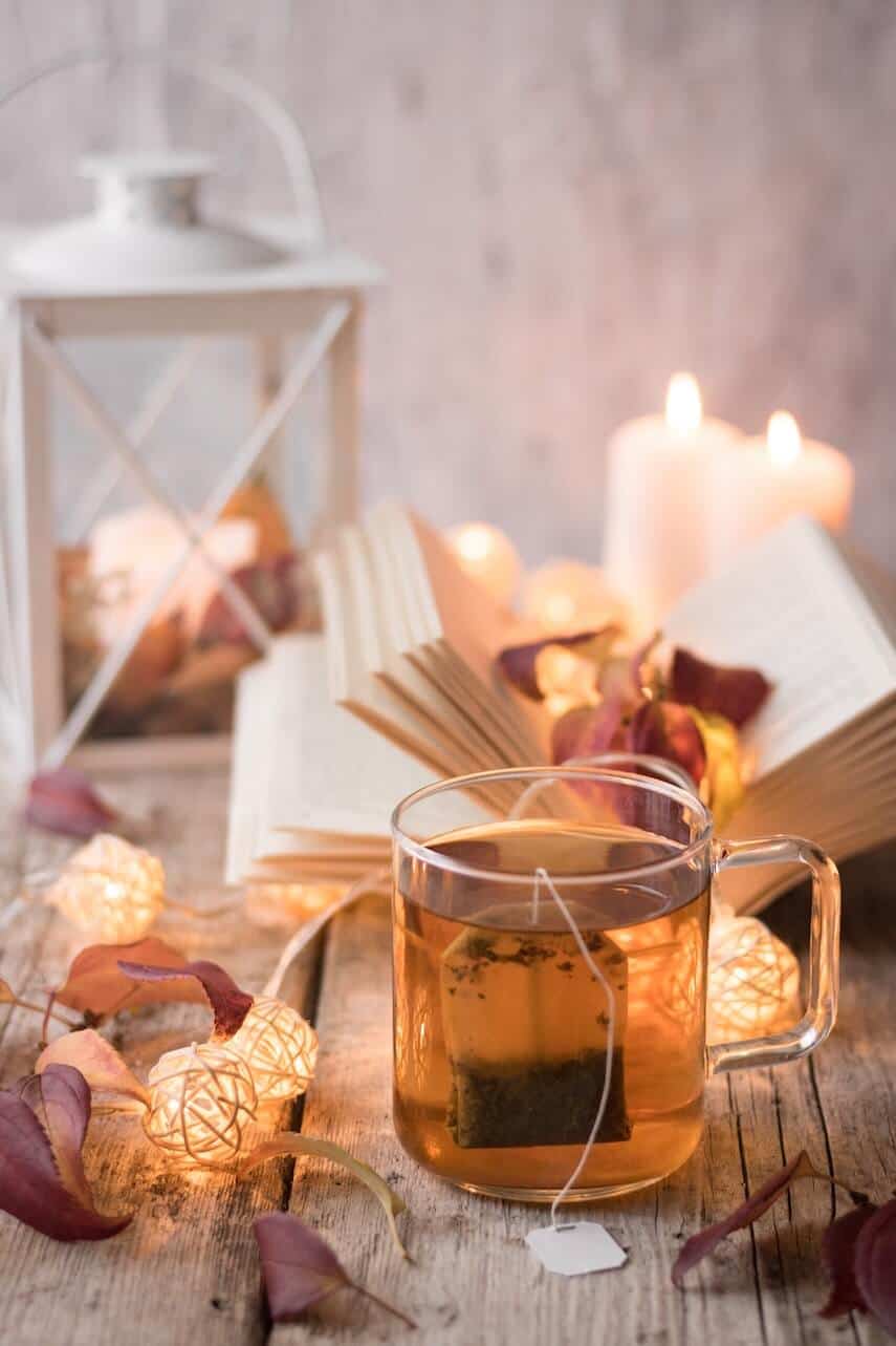 Glass mug of tea with a teabag surounded by wickerball fairy lights in front of an open book and hurricane lantern
