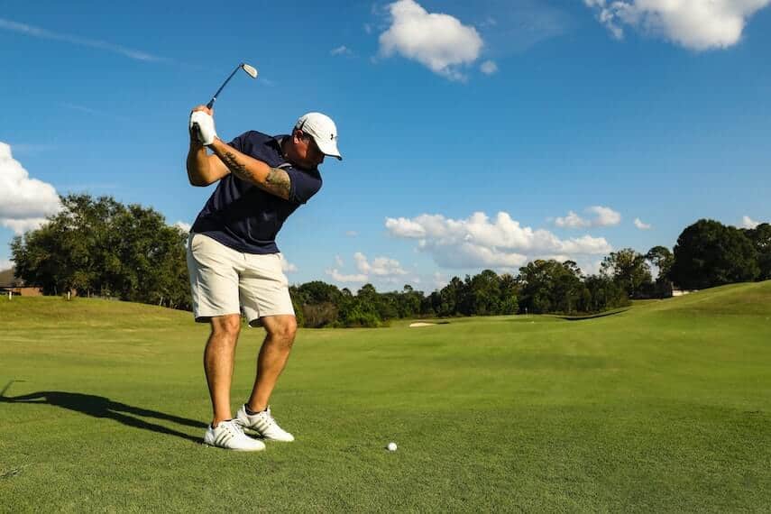 Man in beige shorts and navy shirt taking a backswing, white golf ball on the grass beneath him