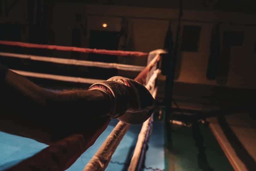 Mans arm holding onto the top rope of a boxing ring while standing in the corner, the far corner out of focus