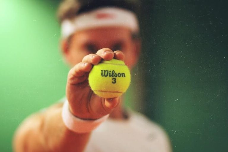 Tennis Quiz Questions and Answers cover photo of an out of focus man earing a white headband holding an in focus tennis ball out in front of his face