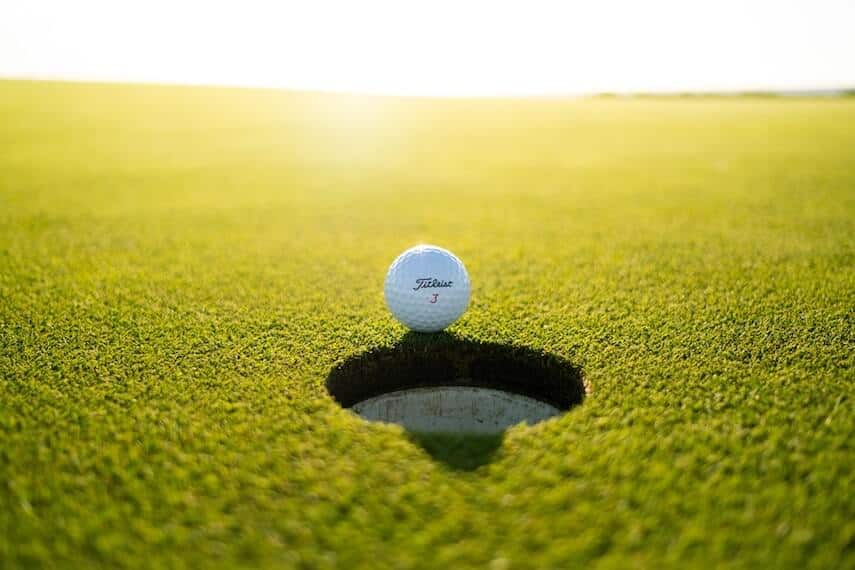 White golf ball sitting at the very edge of the hole, the sun setting behind the shot casting a glow onto the putting green
