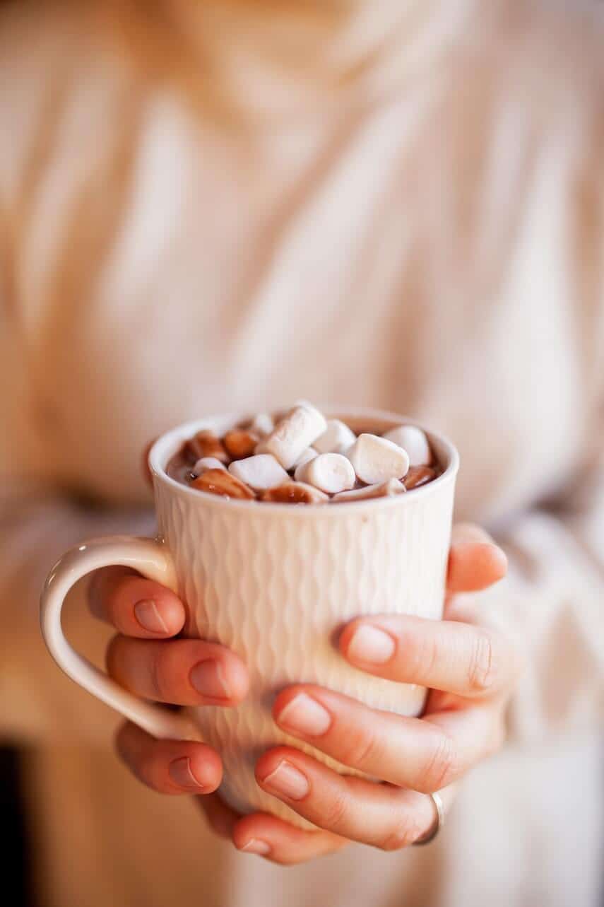 Hands around a white mug of hot chocolate which is topped with small white marshmellows
