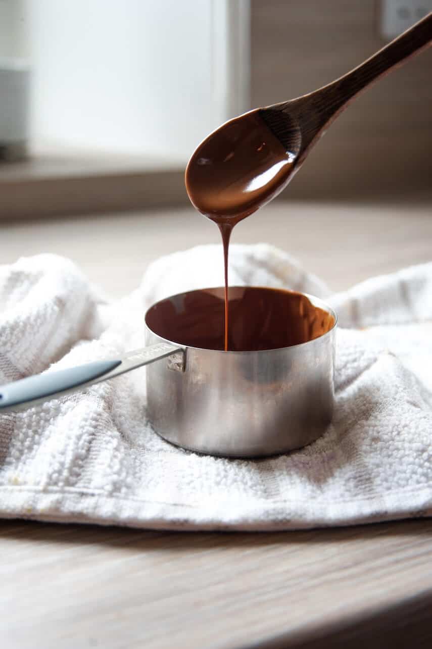 Small silver pot of melted chocolate, a wooden spoon dripping chocolate into the pan