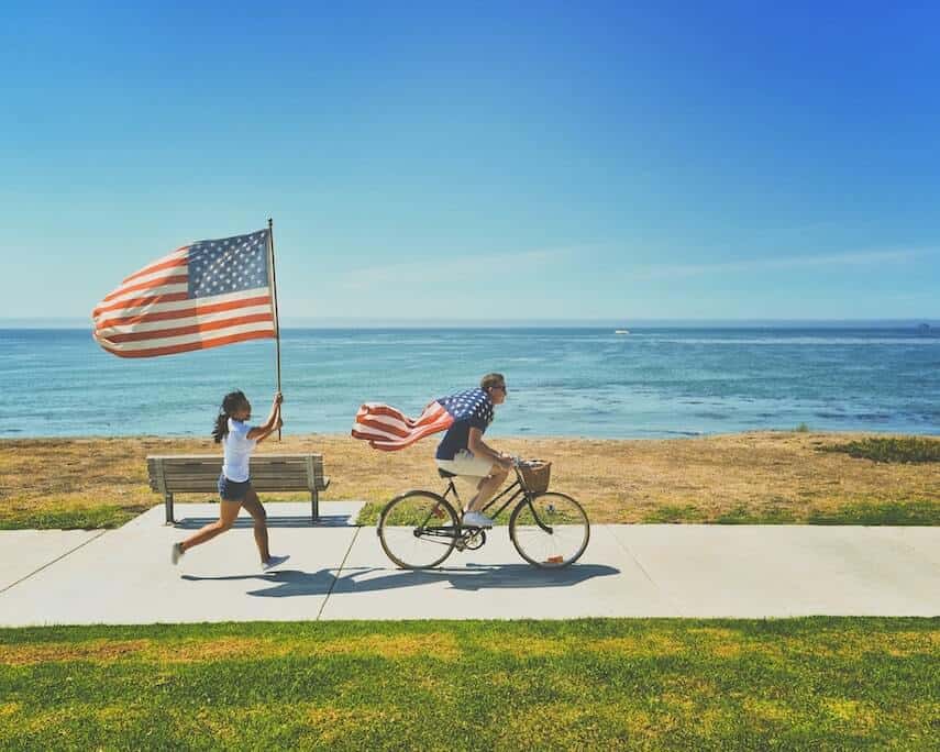 Man riding a bicycle wearing a US flag cape being followed by a woman waving an American Flag