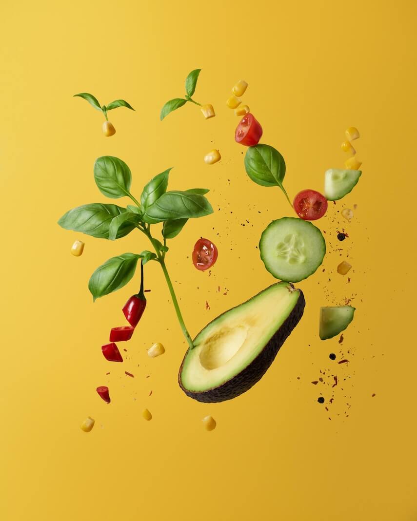Half an avocado floating surrounded by a basil sprig, chopped red chilli, sliced cucumber and halved cherry tomatoes against a yellow background