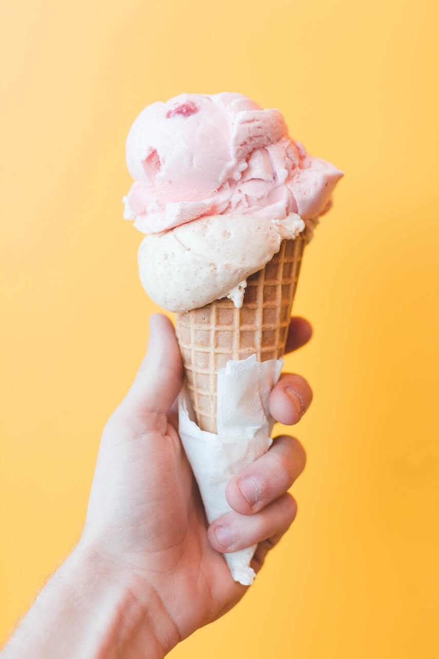 Hand holding an ice cream cone with pink icecream in front of a yellow wall