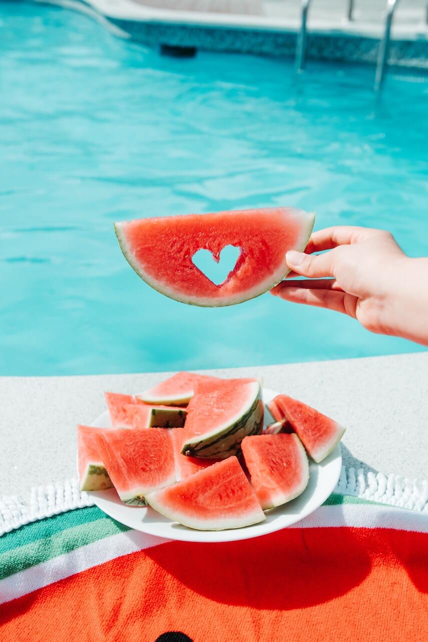 Slices of Watermelon on a plate next to a swimming pool, one slice held up has a heart shape cut out of the flesh