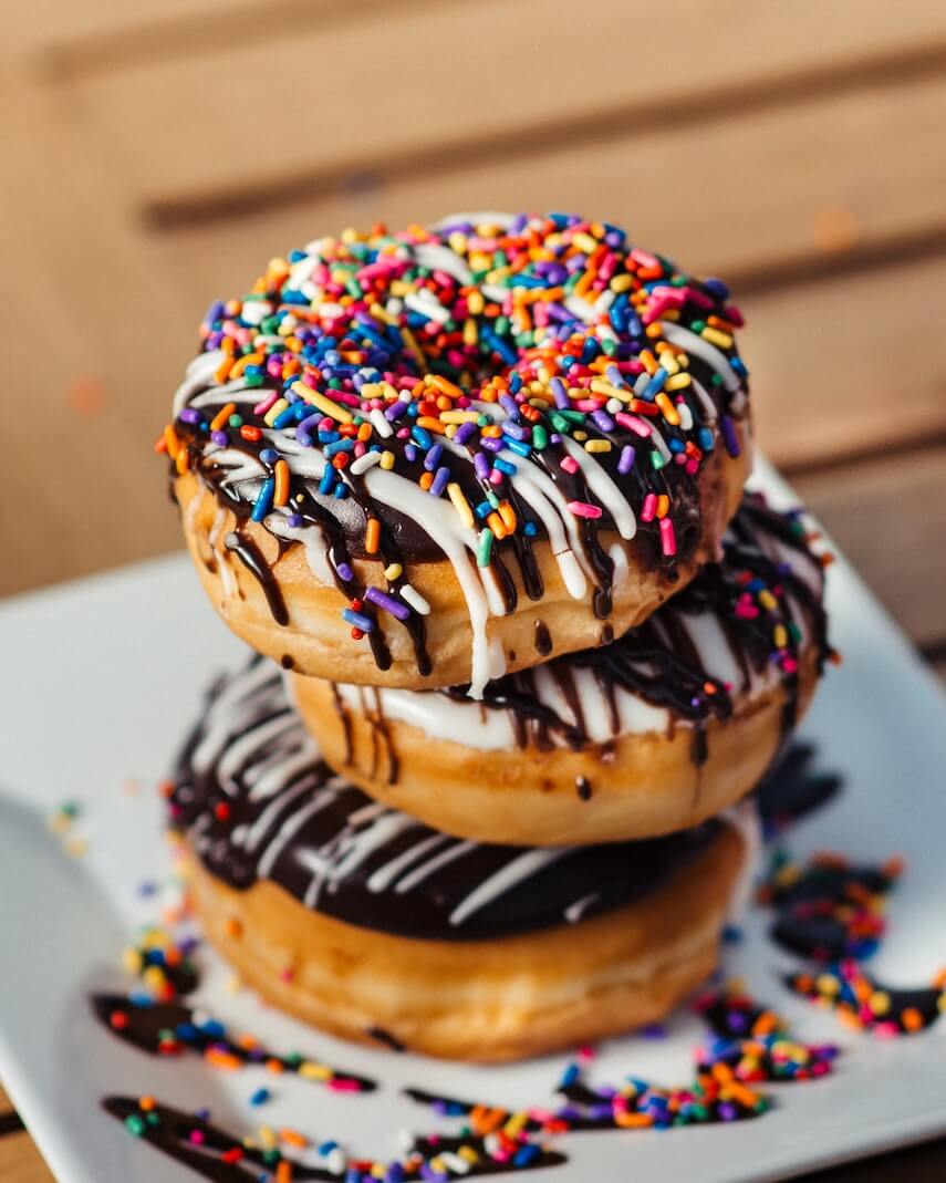 Three chocolate glazed donuts stacked on a plate covered in rainbow sprinkles