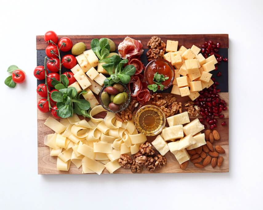 Top down shot of cheese board with blocks of cheese, nuts, pomegranate perls, meats, oil and salsa dips, olives, nuts, tomatoes and basil leaves.