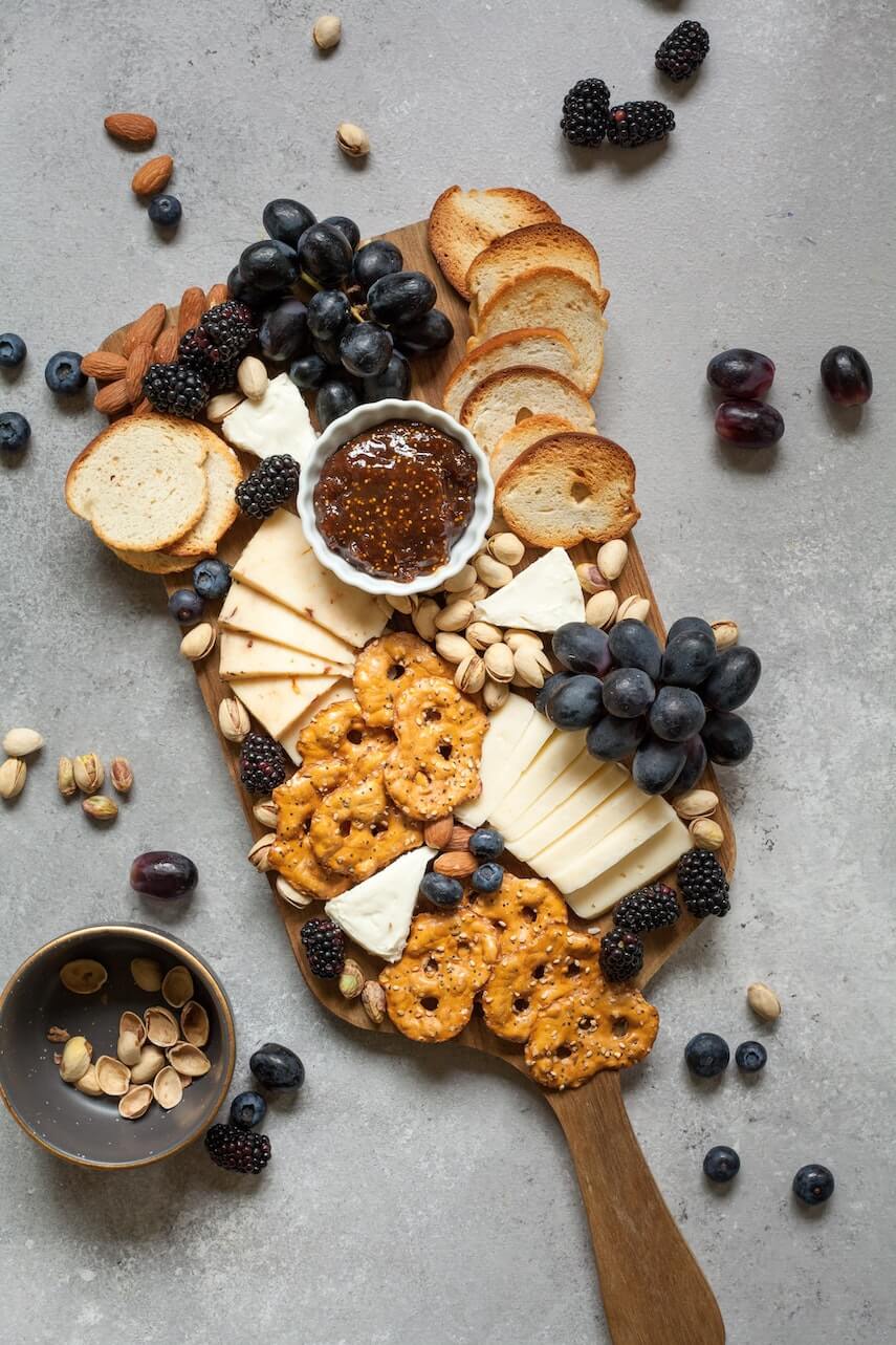 Wooden cheeseboard with cheese, breads, pretzels, grapes, blackberries and dip