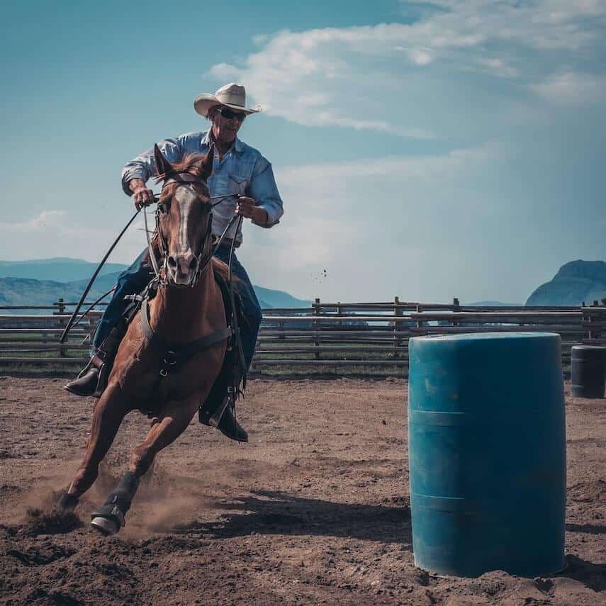 Cowboy barrel racing a brown horse in a dirt paddock