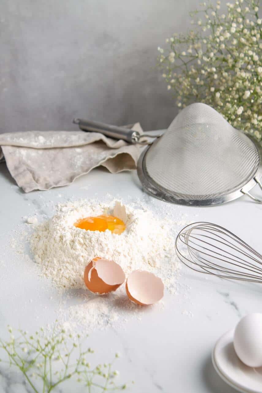 Egg cracked into the well in a mound of flour next to an upturned sieve and silver whisk