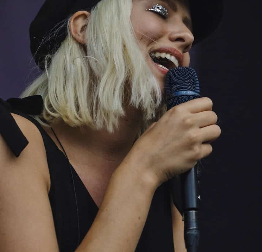 Female with short blonde hair, silver sparkly eyeshaddow and black hat singing into a black microphone