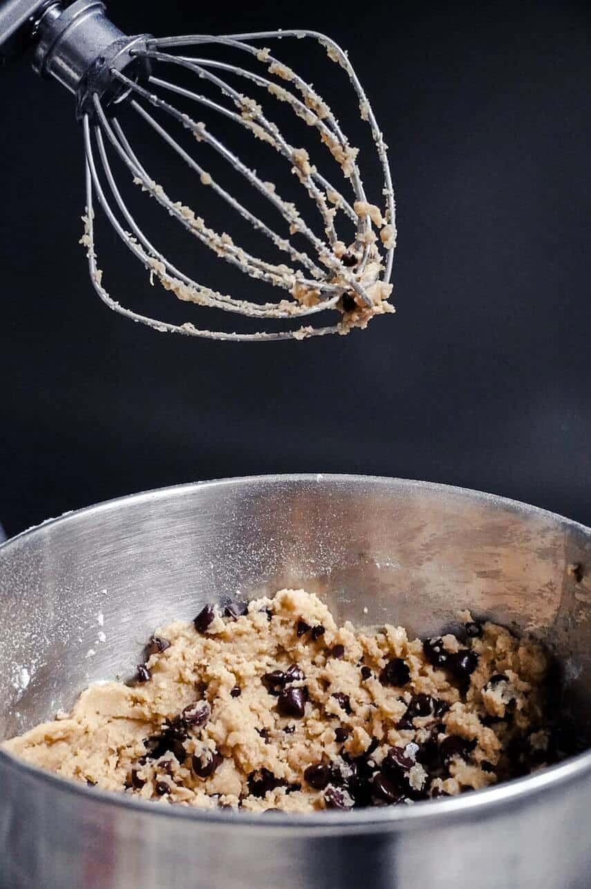 Head of large electric whisk about a silver bowl with dough and chocolate chips in it