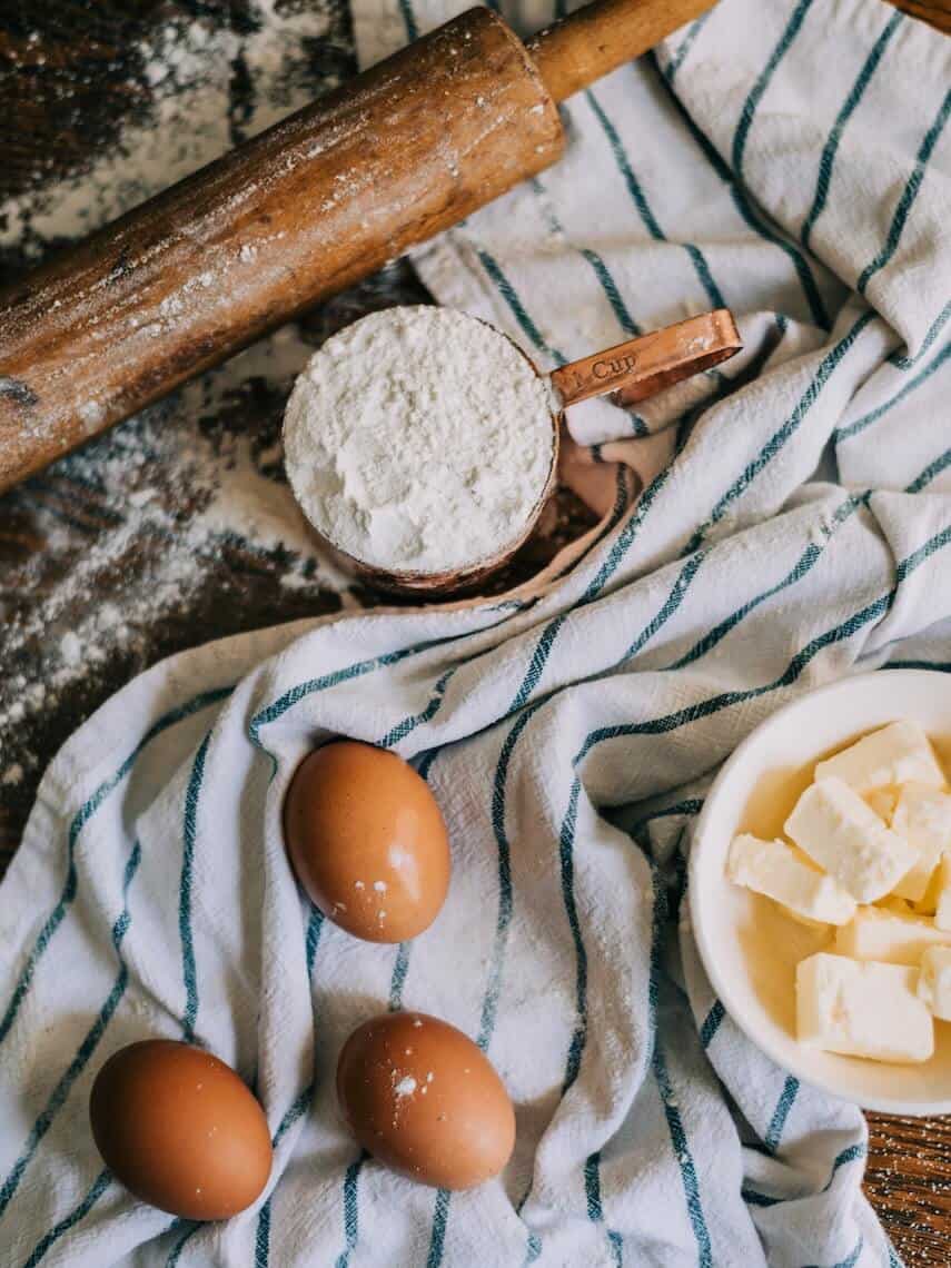 Rolling pin, cup of flour, three eggs and sticks of butter on a striped tea towel