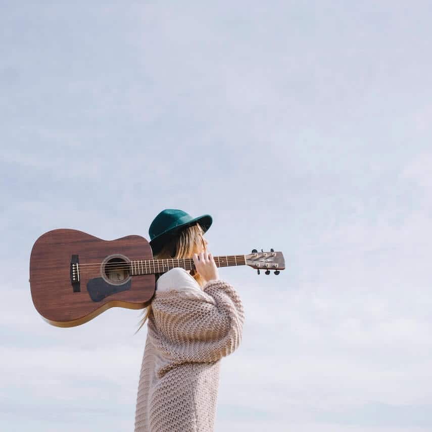 Woman wearing green hat, beige cable knit cardigan standing at a right angle to camera holding the neck of an acoustic guitar on her shoulder