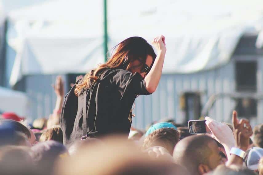 Woman with long brown hair, black top wearing sunglasses on someones shoulders above a crowd of people at a festival