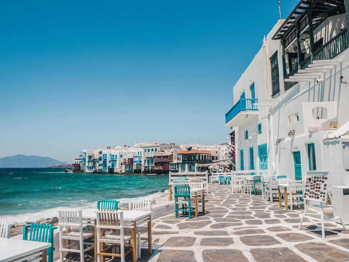 Greece Quiz Cover Photo of White Greek village by the ocean, wooden tables and chairs set up on the cobbled patio