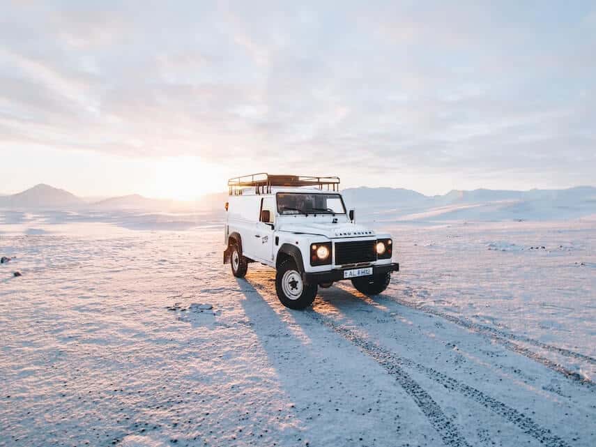 White Land Rover with black roof rack parked on snow, snowy mountains in the distance behind it