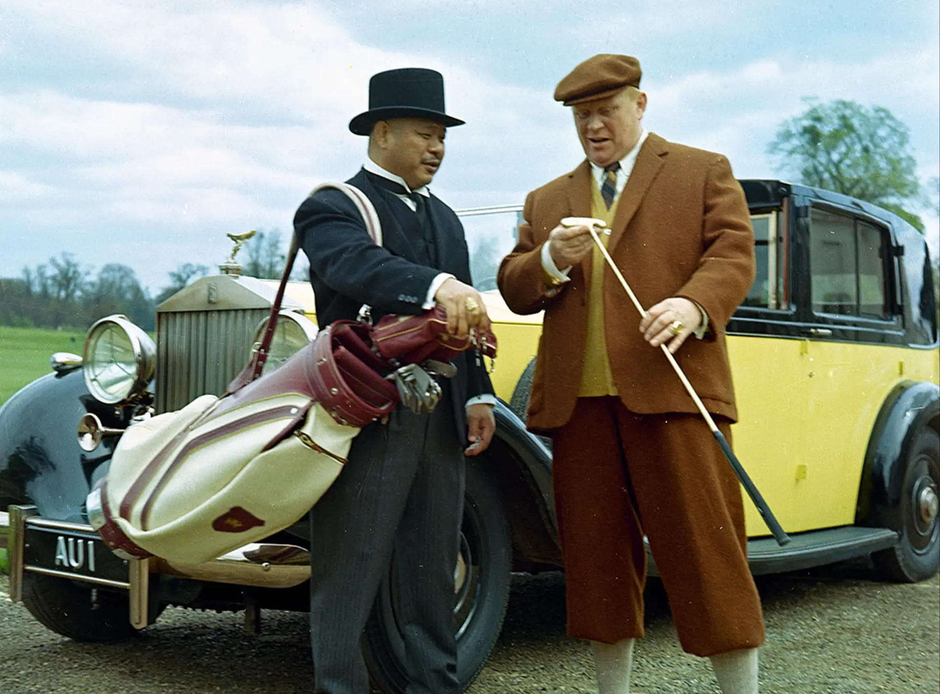 Goldfinger and Oddjob wearing suits holding golf clubs next to a yellow Rolls Royce car