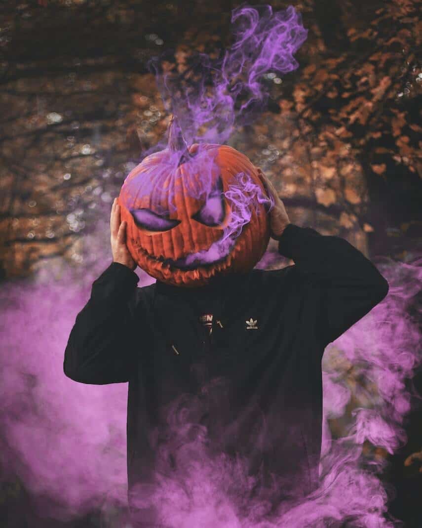 Man wearing a black hoodie with a carved orange pumkin on his head, purple smoke coming from the eyes and mouth