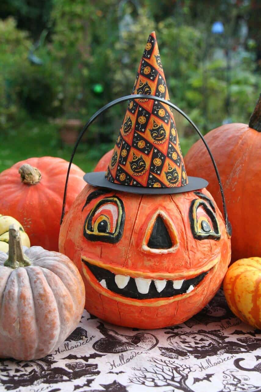 Pumpkin with a goofy face painted on it, wearing a witches hat with balck cats on it