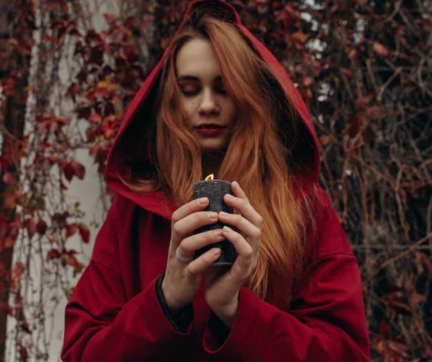Red haired woman holding a black cancle wearing a red hooded coat in a wood