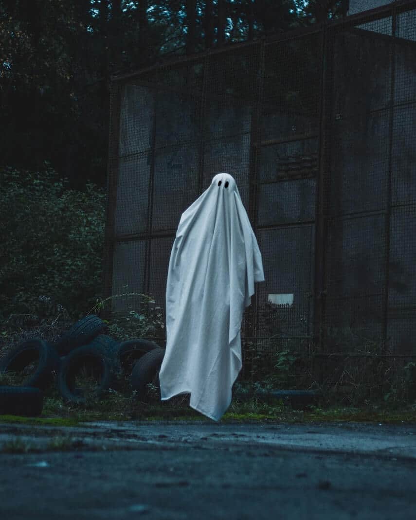 White sheet with eye cut out to look like a ghost, suspended off the floor in front of old tires and a brown cage building