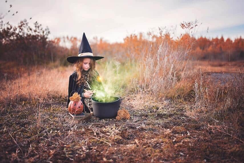 Young girl dressed as a witch holding a wand above a cauldron with green smoke coming out of it, in a clearing in a field